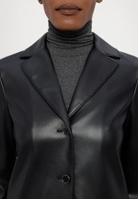 Svart fusk läderjacka med en skarp krage, tvåknappsstängning och slät textur, lagd över en grå polotröja.