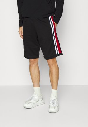 Mann trägt schwarze kniehohe Shorts mit roten und weißen Seitenstreifen, weiße Socken und weiße Sportschuhe, steht vor einem einfarbigen Hintergrund.