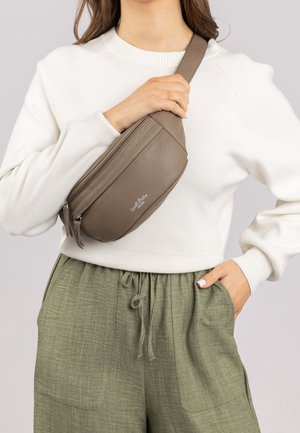 Femme portant un pull blanc et un pantalon vert tient un sac bandoulière en cuir taupe avec des poches zippées sur la poitrine.