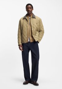 Γκέκα ελιά βαμβακερή με γκρι γιακά, κλείνει με φερμουάρ και κουμπιά με πατίν. Φορεμένη πάνω από μπεζ παλτό, συνδυασμένη με σκούρα τζιν παντελόνια και καφέ παπούτσια.