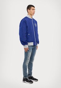 Blue bomber jacket with a gray collar, zip closure, side pockets, and a round logo on the chest; worn with distressed light jeans and black sneakers.