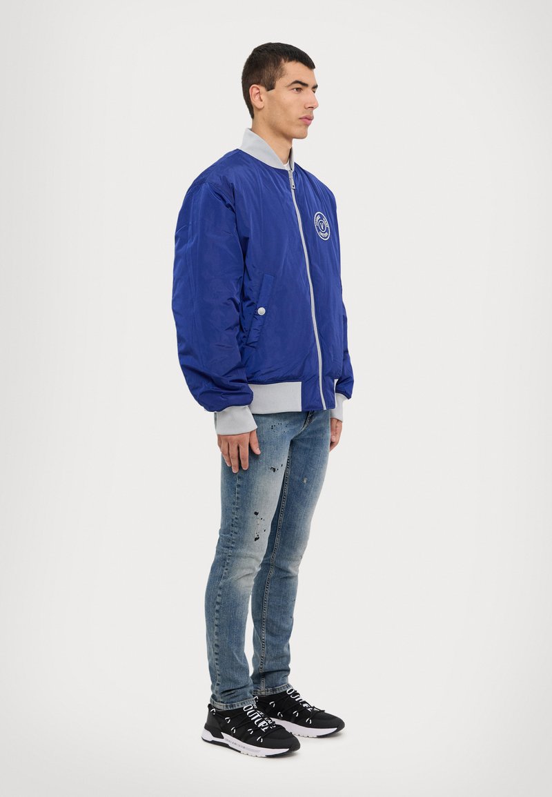 Blue bomber jacket with a gray collar, zip closure, side pockets, and a round logo on the chest; worn with distressed light jeans and black sneakers.