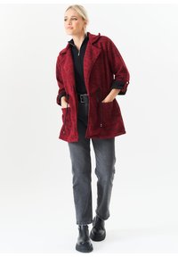 Red textured jacket with a belted waist, large pockets, and a hood, paired with gray jeans and black ankle boots.