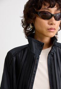 Black pleated jacket with high collar, zip closure, and subtle stitching details, paired with a light beige Nike shirt and black sunglasses.