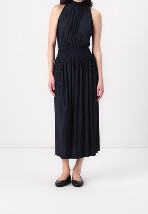 Vrouw die een mouwloze zwarte geplooide midi-jurk met een hoge hals draagt en zwarte balletschoenen, staand tegen een effen achtergrond.