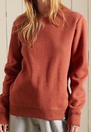 Osoba ubrana w rdzawy sweter z długim rękawem i okrągłym dekoltem, z prążkowanymi mankietami i dolnym ściągaczem, stojąca na tle drewnianych paneli.