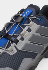 Κοντινή λήψη μπλε και γκρι παπουτσιού πεζοπορίας με ελαστικά κορδόνια γρήγορου τράβήγματος και μαύρο κλείδωμα τάγκλ σε διαπνέον άνω μέρος από δίχτυ.