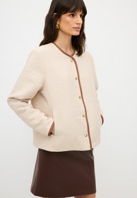 Vadderad beige jacka med bruna kanter, prydd med guldknappar och sidofickor, tillsammans med en brun läder minikjol.