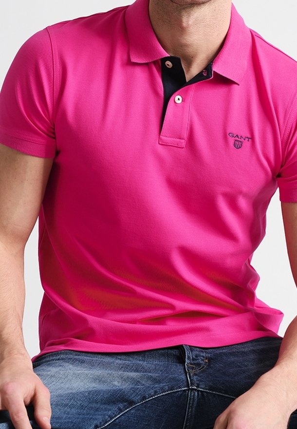 Homme portant un polo rose vif à manches courtes avec un petit logo sur la poitrine et un jean bleu, assis les mains reposant sur les jambes.