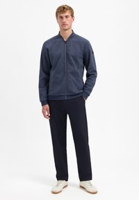 Donkerblauwe zip-up jas met een staande kraag, gestructureerde stof, zakken aan de voorkant en ribgebreide manchetten, gecombineerd met marineblauwe broeken en lichte sneakers.