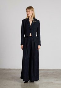 Vrouw gekleed in een zwart gestreepte cropped blazer en wijdvallende high-waisted broek, staand op een betonvloer tegen een effen muur.