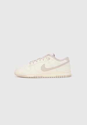 Basket basse beige Nike avec logo Swoosh beige clair, lacets et semelle texturée sur un fond gris clair uni.