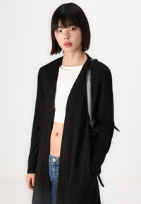 Black long cardigan made of soft material, open front, with side pockets. Worn over a white crop top and paired with blue jeans.