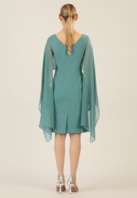 Türkises Kleid mit durchsichtigen Cape-Ärmeln, V-Ausschnitt, taillierter Rückseite, knielanger Länge und einem Mittel-Schlitz; kombiniert mit metallischen Absatzschuhen.