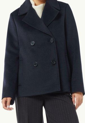 Kobieta ubrana w granatowy dwurzędowy płaszcz typu pea coat, pod którym ma biały dzianinowy sweter i czarne spodnie w prążki, stojąca z rękami swobodnie opuszczonymi.