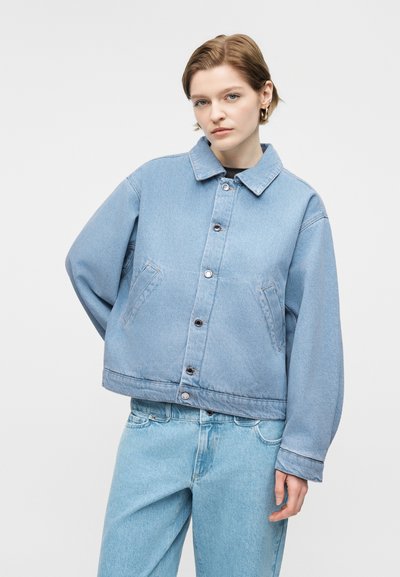 Νεαρό άτομο με κοντά μαλλιά φοράει μια ανοιχτό μπλε υπερμεγέθη τζιν μπουφάν και ανοιχτό μπλε τζιν παντελόνι, στέκεται μπροστά σε απλό γκρι φόντο.