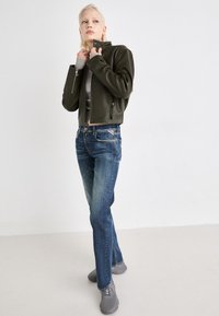 Chaqueta corta de cuero verde oscuro con bolsillos con cremallera, combinada con jeans ajustados azules y zapatillas grises. Diseño simple y elegante con acentos mínimos.