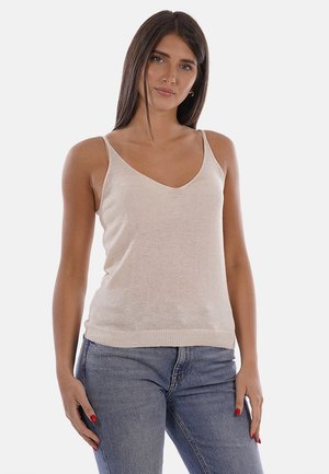 Mujer con cabello largo y castaño lleva una camiseta sin mangas beige con cuello en V y vaqueros azules, de pie frente a un fondo claro y sencillo.