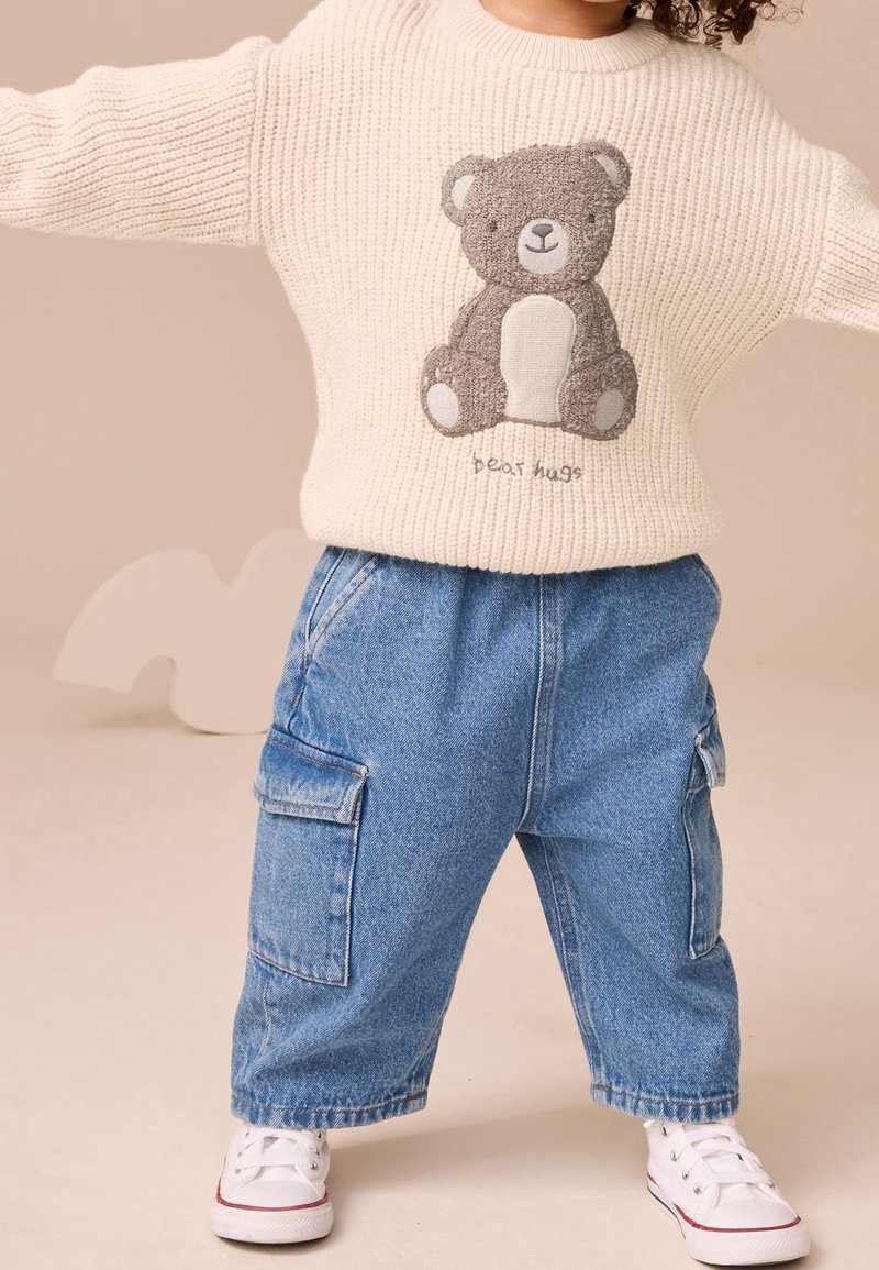 Enfant portant un pull en maille crème avec un ours en peluche et le texte « bear hugs », jean cargo bleu ample et baskets blanches, debout les bras écartés.