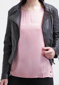 Schwarze Lederjacke mit gesteppten Schulterakzenten, kombiniert mit einer hellrosa Bluse. Die Jacke hat ein tailliertes Design und silberne Reißverschlüsse.