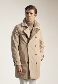 Abrigo beige de doble botonadura con grandes botones oscuros, solapas anchas y un ajuste cómodo. Se lleva sobre un suéter de cuello alto acanalado.