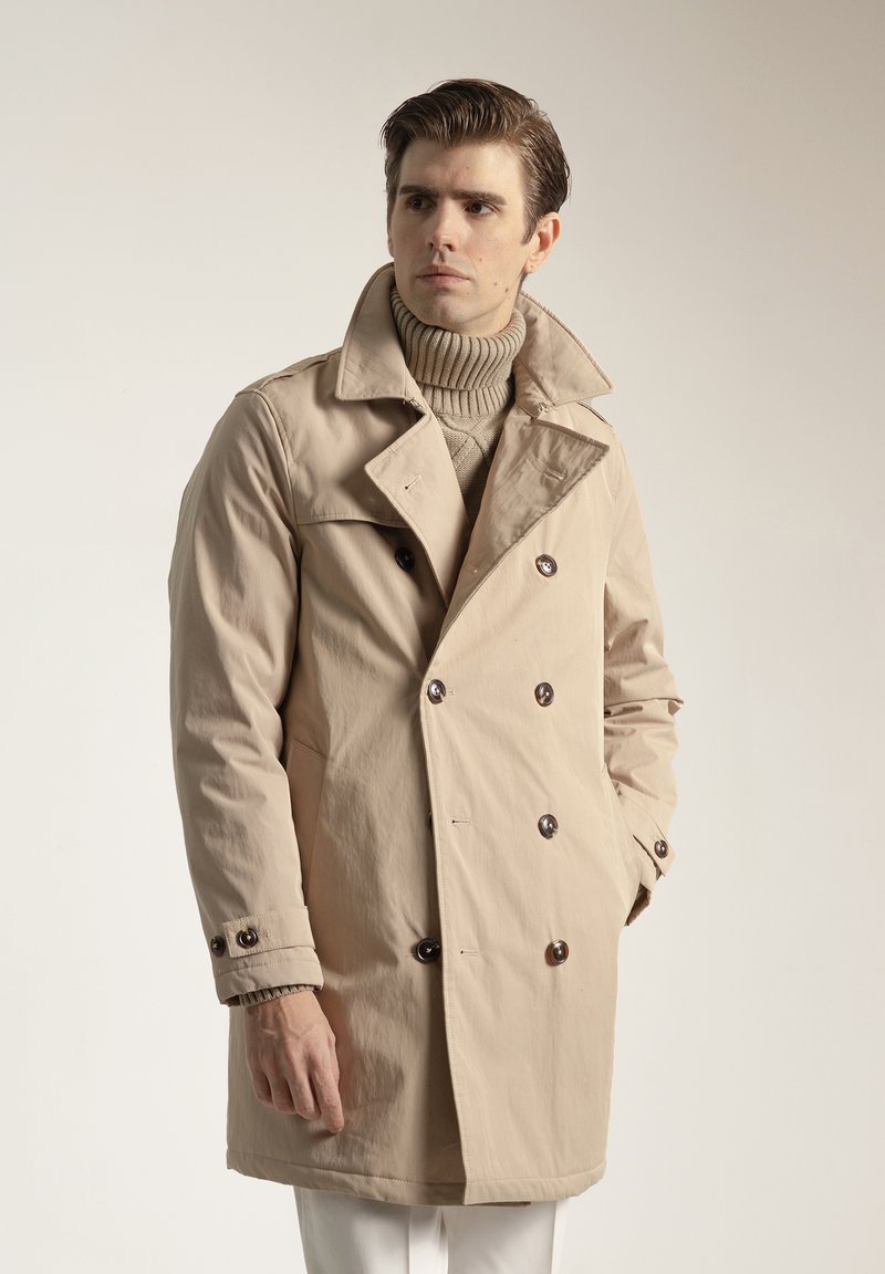 Abrigo beige de doble botonadura con grandes botones oscuros, solapas anchas y un ajuste cómodo. Se lleva sobre un suéter de cuello alto acanalado.