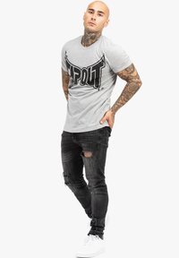 Pelēks īsām piedurknēm T-krekls ar melnu "TAPOUT" logotipu, saskaņots ar nolietotām melnām džinsām un baltām sporta apaviem. Modelis stāv ar vieglu slīpumu.