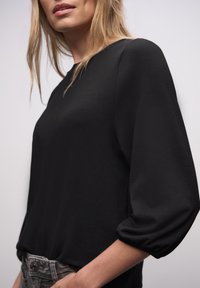 Czarna, długorękawowa bluzka o luźnym kroju z marszczonymi mankietami. Gładka faktura materiału, elegancki dekolt i minimalistyczne detale.