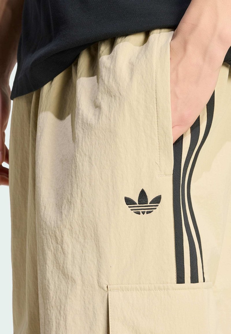 Beige Shorts aus strukturiertem Stoff mit Seitentaschen und schwarzen drei Streifen als Akzent. Auf dem linken Oberschenkel befindet sich das schwarze Adidas-Logo.
