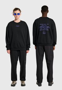 Dwóch modeli nosi czarne bluzy i spodnie; z przodu bluza ma logo "HALO", z tyłu znajdują się niebieskie napisy z terminami taktycznymi i szkoleniowymi.