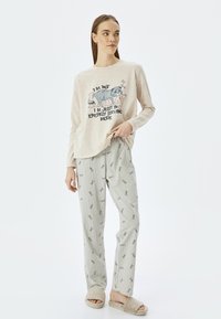 Beige pitkähihoinen paita, jossa on kissakuva ja teksti; raidalliset pyjamahousut, joissa on toistuva vaaleanharmaa teksti; pörröiset beige tossut; rento malli.