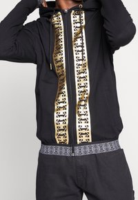 Sudadera con cremallera negra que presenta una franja gráfica dorada en el centro, con una cinturilla con patrón. Tejido suave y cordones ajustables.