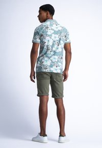 Lyhythihainen vaaleansininen kukallinen poolopaita, jossa on vihreitä ja valkoisia kukkia. Yhdistettynä kyynärpäitä ylös käärittyihin oliivinvihreisiin shortseihin ja valkoisiin lenkkareihin.