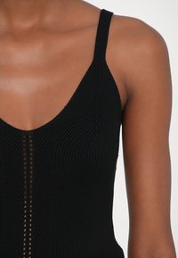 Černý žebrovaný pletený tílko s hlubokým výstřihem do V, úzkými ramínky a svislou linií malých výřezů.