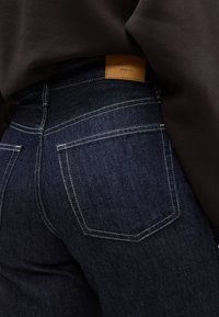 Sötétkék farmernadrág magas derékkal, barna bőr címkével és kontrasztos fehér varrással a hátsó zsebeknél.