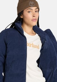 Marineblå kordfløjljakke med lynlås, med høj krave og forlommer, parret med en lys cremefarvet skjorte med et orange logo.