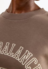 Brown sweatshirt with a ribbed crew neckline, featuring white lettering design that reads "BALANCE" across the chest. Smooth fabric texture.