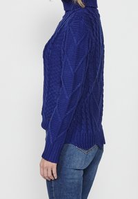 Pull à col roulé en maille torsadée bleu roi. Présente des motifs texturés et une coupe décontractée. Poignets et ourlet côtelés avec des fentes latérales.