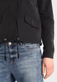 Svart vindjacka med två framfickor, justerbara dragsnören i midjan och en mjuk textur, i kombination med slitna blå jeans.