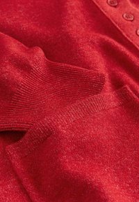 Pull-over rouge en tricot doux. Comprend des poignets côtelés et une patte de boutonnage. Un léger éclat dans le fil ajoute de la texture.