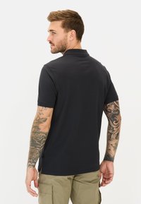 Marineblå polo-shirt med korte ærmer, sideskår og klassisk krave, båret med beige cargobukser. Har komplekse tatoveringer på armene.