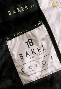 Intérieur d'une veste en fausse fourrure noire avec une étiquette crème. L'étiquette affiche le nom de la marque, les instructions d'entretien et des icônes pour la résistance à l'eau et la chaleur.