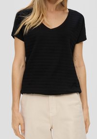 Žena oblečená v černém krátkém rukávu s výstřihem do V a texturovaném topu, s béžovými kalhotami, stojící s uvolněnýma rukama podél těla.