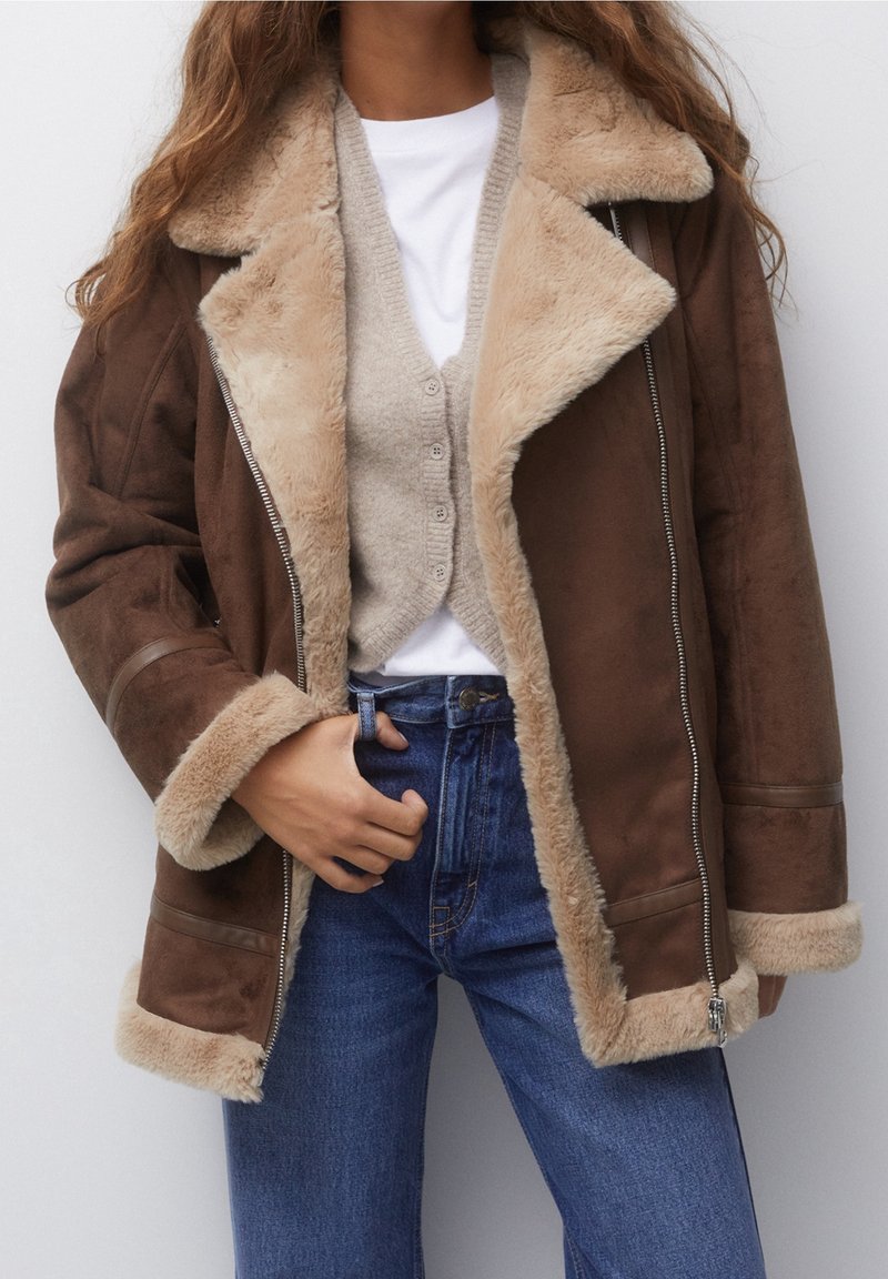 Brun shearling jakke med beige pelsfor, båret over en beige cardigan og hvid skjorte, parret med højtaljede blå jeans.