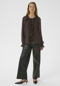 Naine kannab läbipaistvat tumepruuni volangiga pluusi, musti laiadest säärest nahkpükse ja musti terava ninaga slingback kingi, seistes valgel taustal.