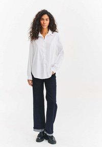 Biała koszula na guziki z długimi rękawami, o luźnym kroju i małym logo na kołnierzyku, zestawiona z ciemnoniebieskimi szerokimi dżinsami i czarnymi butami.