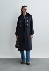 Dunkelblauer Wollmantel mit doppelter Knopfleiste, Toggle-Verschlüssen, Seitentaschen und einem Kunstfellkragen, kombiniert mit hochtaillierten Jeans.