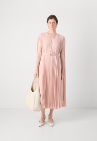 Pleated, blush pink dress with a deep V-neck, belted waist, and cape-like sleeves. Paired with a white bag and pointed beige heels.