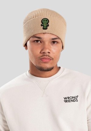 Man in een beige gebreide beanie met een groen gestikt poppatch en een witte sweatshirt met het "WRONG FRIENDS" logo op een lichtgrijze achtergrond.