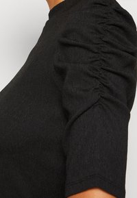 Černý top s texturovaným materiálem, krátkými rukávy s nařasenými detaily a klasickým kulatým výstřihem. Detailní pohled zvýrazňuje design rukávu.